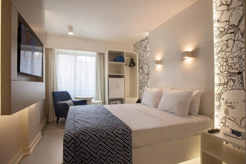 Quarto com uma cama de casal, tv na parede, cadeira azul, janela com cortina branca, frigobar e paredes brancas com detalhes pretos, ilustrando post hotéis Ibis no Rio de Janeiro.