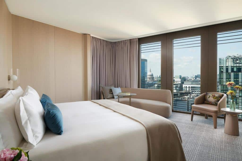 Quarto do Pan Pacific London com cama de casal, janelas panorâmicas, um sofá com almofadas e uma poltrona, o carpete é cinza e branco