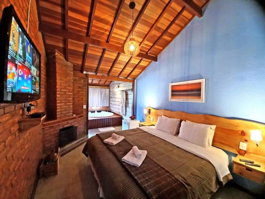 Quarto com cama de casal, parede azul, banheira ao fundo, lareira e TV na parede de tijolos.