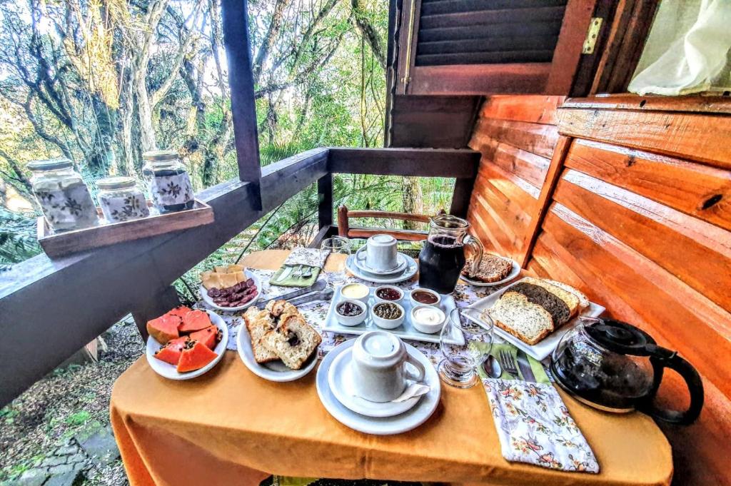 uma mesa de café da manhã farta, com café, suco, pães, frutas, bolos e geleias. A mesa está na varanda do chalé, com vista para as árvores da região.