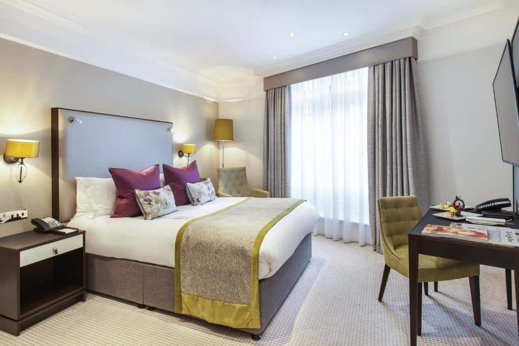QUarto do St. James' Court, A Taj Hotel, London com uma janela com cortinas, cama de casal, uma mesa com uma poltrona, uma televisão, duas mesas de cabeceira com abajures, para representar hotéis bem localizados em Londres