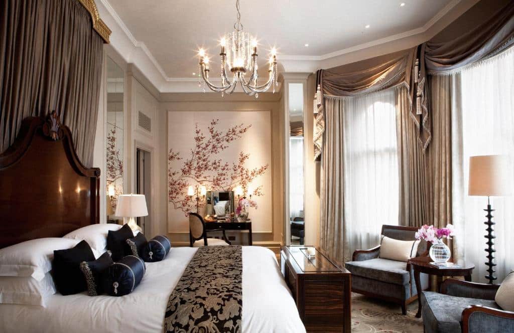 Quarto luxuoso do The Langham London com janelas com cortinas, um lustre, uma cama de casal com muitas almofadas e travesseiros, uma penteadeira, duas poltronas e um abajur de chão