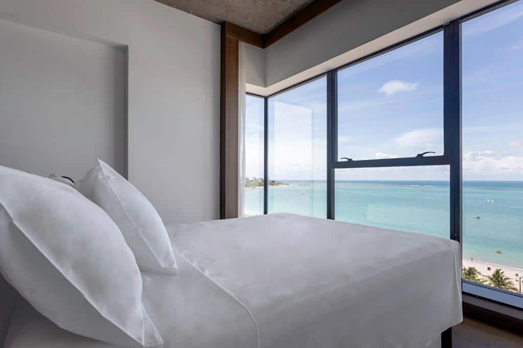 Quarto Duplo do Ibis styles Maceió, de 17 m², com uma cama de casal em frente à uma parede de vidro com vista panorâmica do mar. Representa hotéis em Maceió
