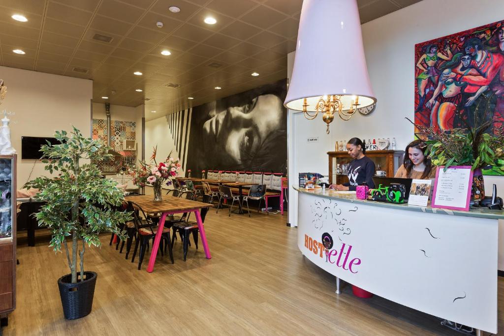 Recepção do Hostelle, um hostel só para mulheres em Amsterdam, com um balcão escrito "Hostelle" e duas mulheres sorrindo atrás e, ao fundo da imagem, tem mesas e cadeiras, além de flores e um desenho preto e branco do rosto de uma mulher em uma parede enfeitando o espaço