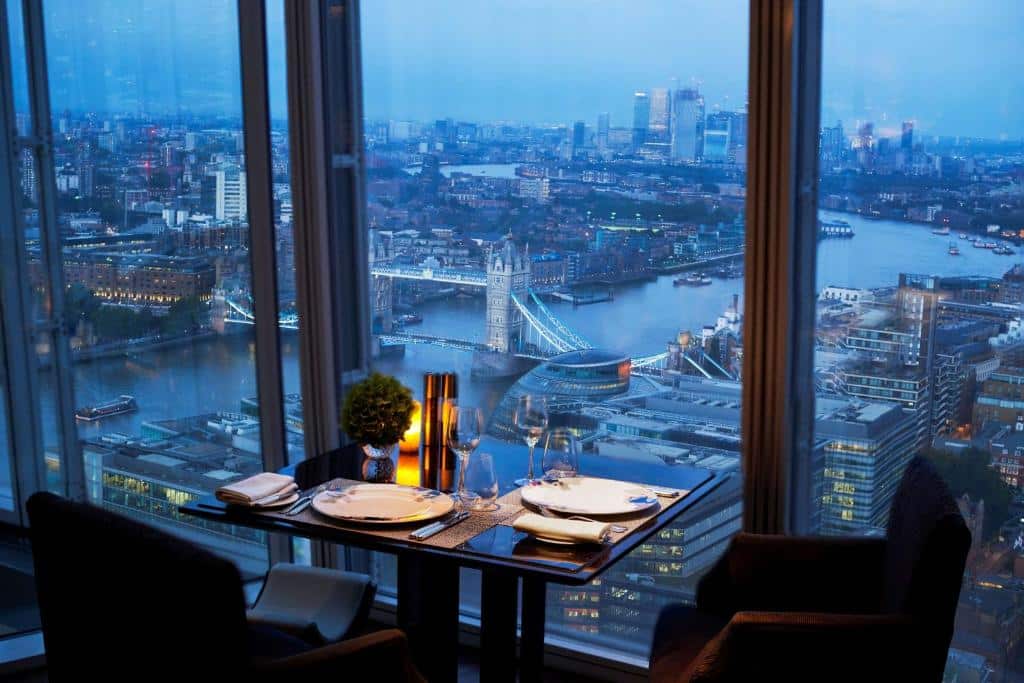 Área de refeições do Shangri-La The Shard, London com uma janela panorâmica dando vista para a cidade, há uma pequena mesa com dois lugares, para representar os melhores hotéis em Londres