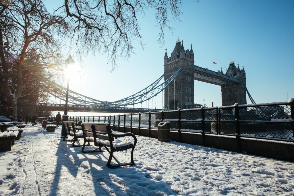 Tower Bridge coberta por neve com bancos e uma grade beirando o rio com postes de luz e árvores secas, a foto tirada no inverno e é possível enxergar toda a ponte