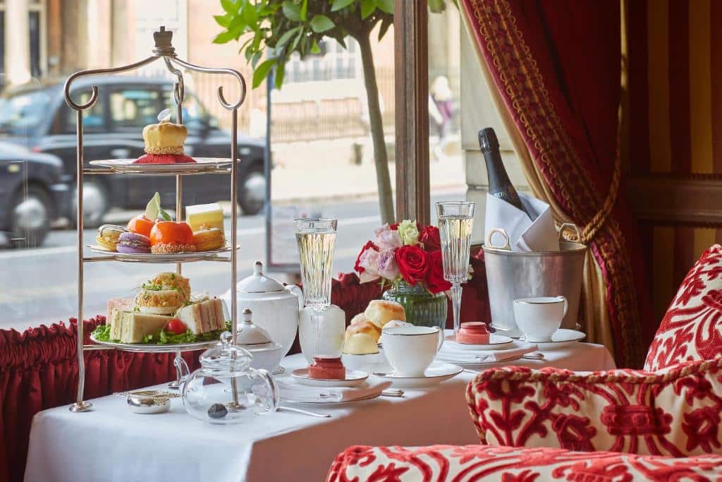 Área de refeições do Rubens At The Palace com uma mesa para duas pessoas com champanhe, vaso de flores, doces e pãezinhos, com vista para a rua, para representar hotéis românticos em Londres
