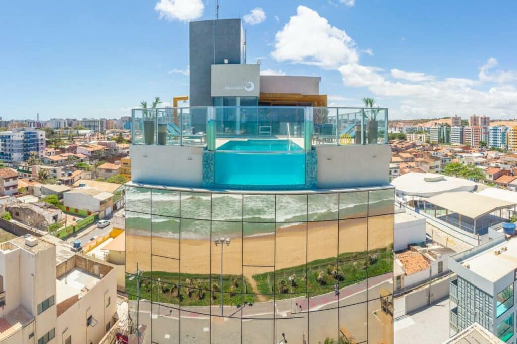 Prédio espelhado do hotel Água de Coco, repletindo o mar no espelho, e na sacada tem uma área de lazer com uma piscina de borda infinita