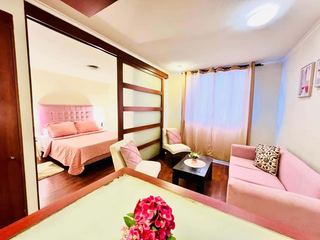 Sala do Infinity Apartments (Huérfanos y Merced) com sofá rosa do lado direito no centro uma mesa e duas poltronas brancas em frente ao sofá rosa. No outro cômodo há uma cama de casal com cobertas rosa. Representa Santiago.