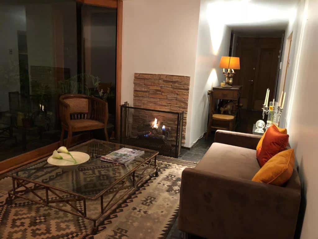 Sala do Rent apart Las Condes com sofá marrom claro do lado direito, mesa de vidro no centro da sala perto de uma lareira.