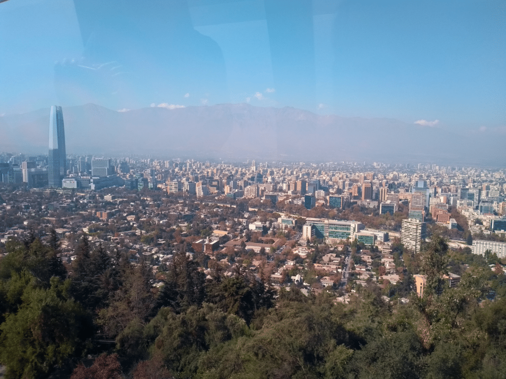 Vista de cima do Funicular do Cerro San Critóbal, em Santiago, durante o dia com prédios em volta e ao fundo as montanhas chilenas. Representa Santiago.