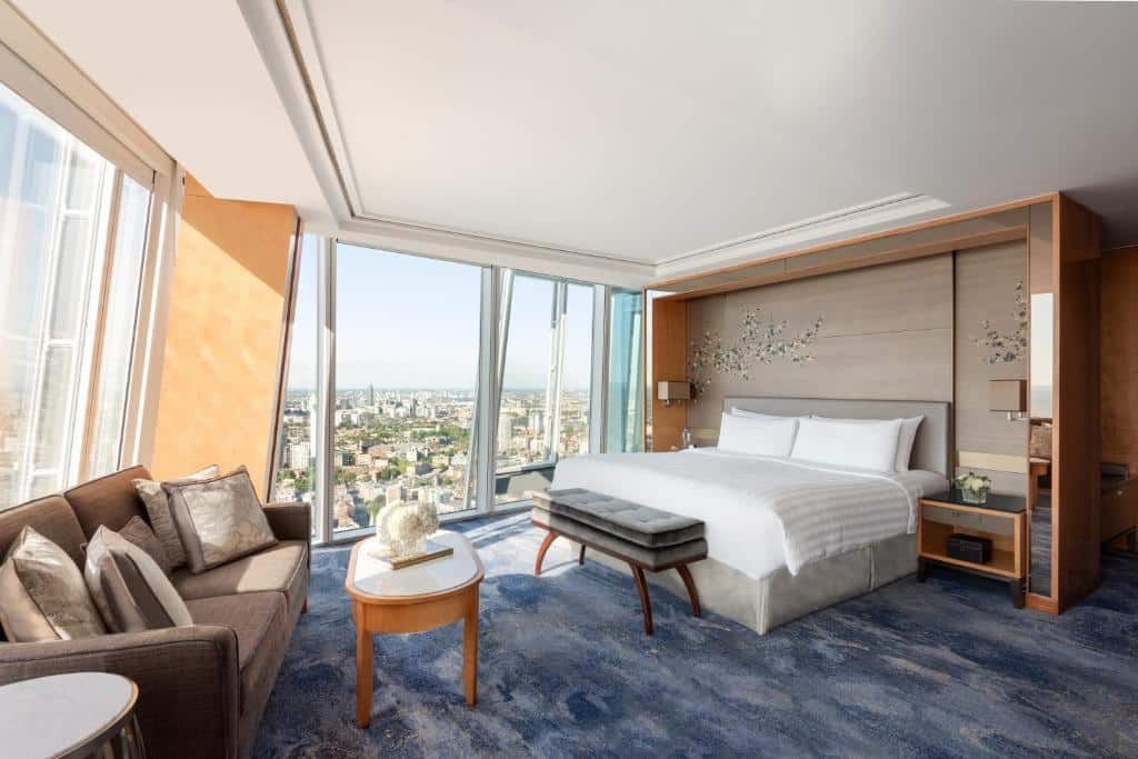 Quarto amplo do Shangri-La The Shard, London com janelas panorâmicas em todo o ambiente, com uma cama de casal, um sofá com almofadas e carpete azul com detalhes em branco