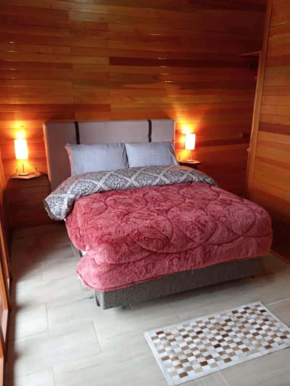 o quarto da Cabana do Lago com vista para Lago São Bernardo é feito em madeira e conta com uma cama de casal no centro do espaço. Há dois abajures, um de cada lado da cama.