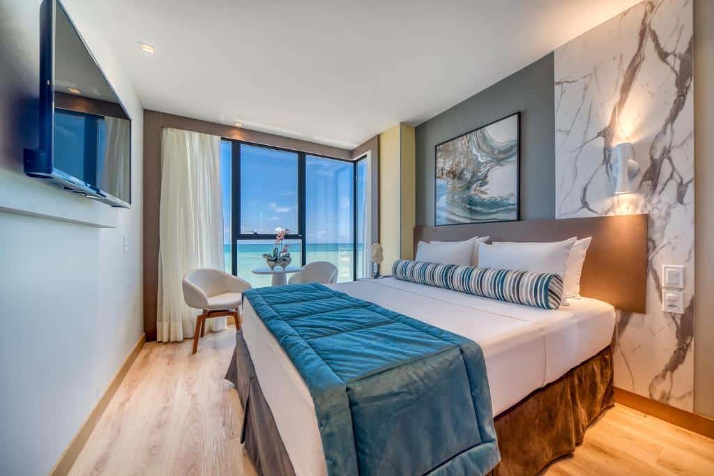 Suíte Premium do Hotel Água de Coco, de 17 m², com uma cama de casal, TV suspensa na parede à frente, e uma mesa com duas cadeiras estofadas e uma janela com vista panorâmica do mar. Representa hotéis em Maceió