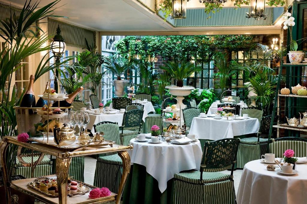 Área de refeições do The Chesterfield Mayfair com tudo decorado em tons de verde, muitas plantas espalhadas pelo local e presas no teto junto de luminárias, as mesas são redondas com cadeiras de madeira verde, para representar os hotéis românticos em Londres