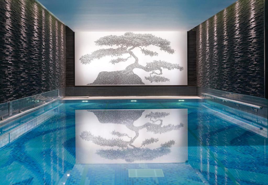 Uma piscina coberta no The Langham London com paredes em pedras pretas e um mosaico desenhado na parede de frente com a piscina, gerando um reflexo da árvore dentro da água