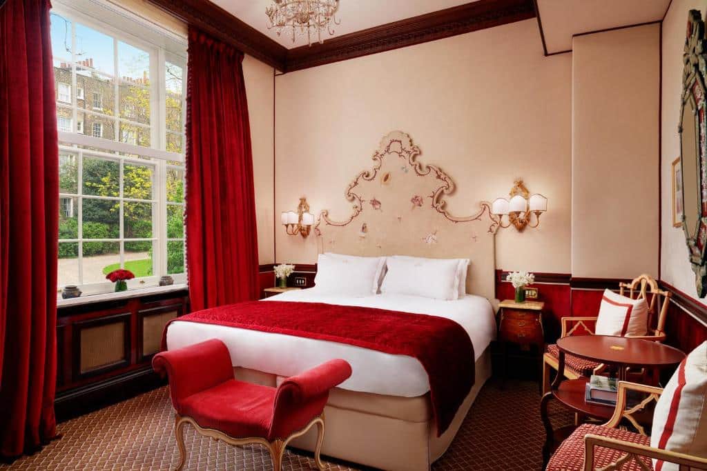 Quarto do The Montague On The Gardens com uma cama de casal, uma janela bem ampla com cortinas vermelhas, uma mesinha com duas cadeiras, o carpete é vermelho e branco, com lustres presos na cabeceira, tudo em estilo bem europeu, para representar os melhores hotéis em Londres