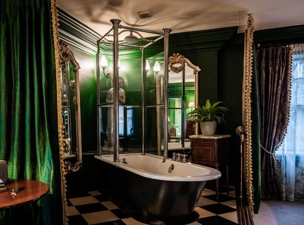 Banheiro amplo do The Rookery  com uma banheira oval, há cortinas verdes e o chão é branco e preto, tudo em estilo europeu e um móvel de madeira com um vaso de flor