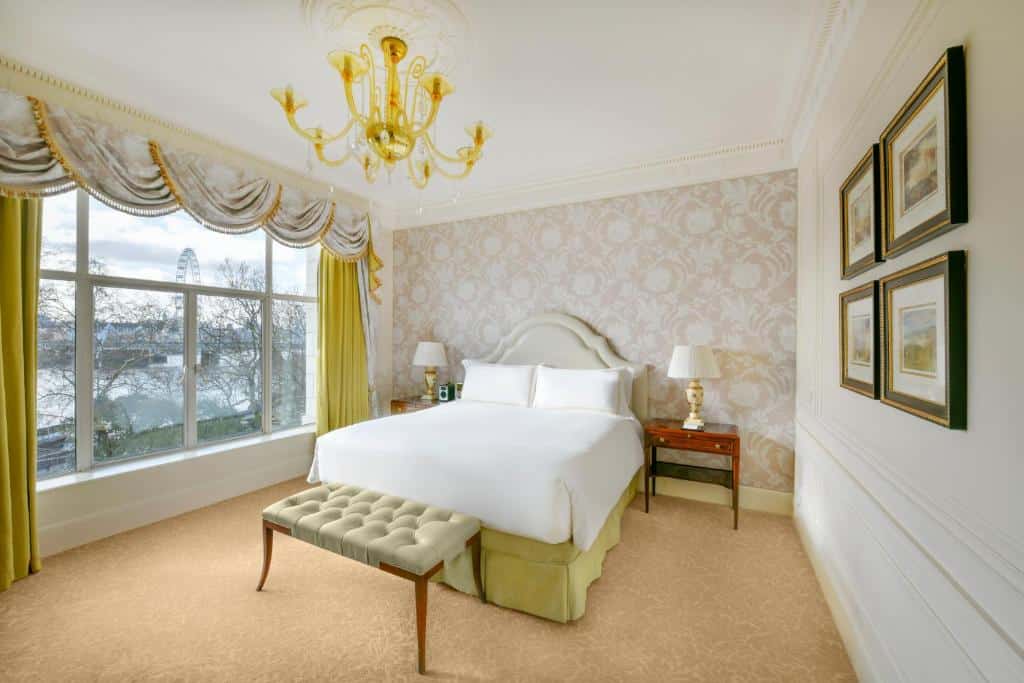 Quarto do The Savoy com uma janela ampla com cortinas, cama de casal, um lustre, carpete bege, alguns quadros na parede e dois abajures