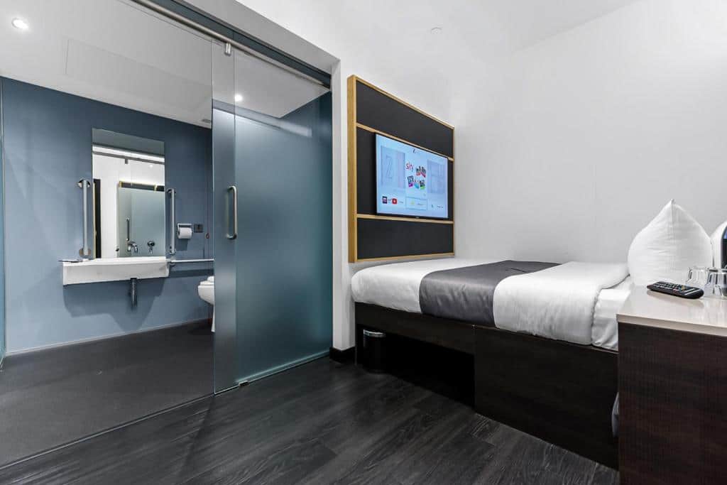 Quarto do The Z Hotel Trafalgar com uma cama de casal e uma televisão em frente, uma mesa de cabeceira, e uma porta de vidro que leva ao banheiro, para representar hotéis perto da London Eye em Londres