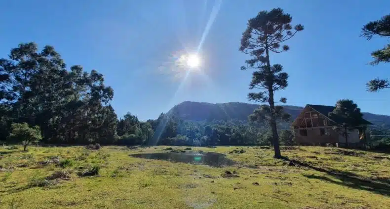 área aberta e externa da Cabana 16, uma entre os chalés em São Francisco de Paula. Há uma pequena cabana de madeira no canto direito da imagem, no restante é possível ver bastante gramado e árvores verdes, além de um céu azul e ensolarado