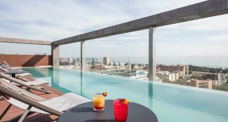 Piscina do Acta Voraport, uma das recomendações de hotéis baratos em Barcelona. Dois drinks coloridos estão em cima de uma mesinha ao lado de espreguiçadeiras. Há uma balaustrada de vidro com vista para a cidade.