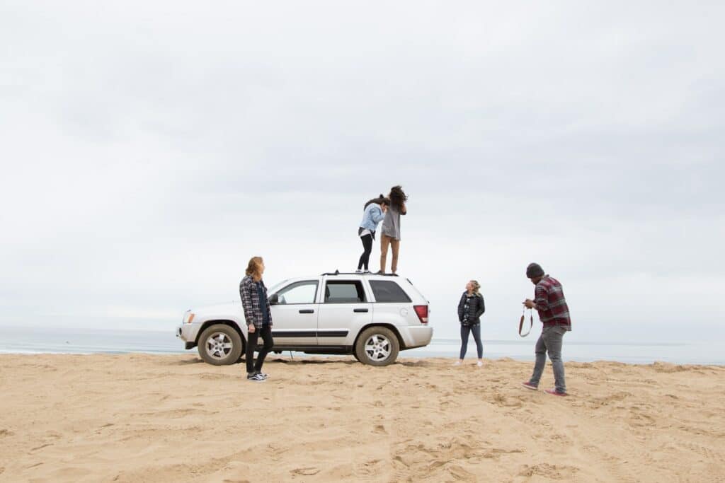 No centro da foto há um carro prateado, com duas garotas em cima dele, no teto. Ao redor, há três pessoas paradas, duas observando as garotas, e um olhando uma câmera na mão. O chão é de areia e ao fundo o mar se mistura com o céu, num dia nublado.