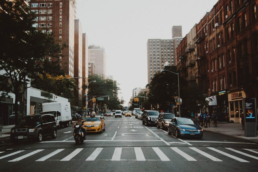 Rua de Nova York, com carros parados, moto e táxi, faixa de pedestres vazia em frente, e prédios em ambos os lados, para ilustrar o post sobre aluguel de carros