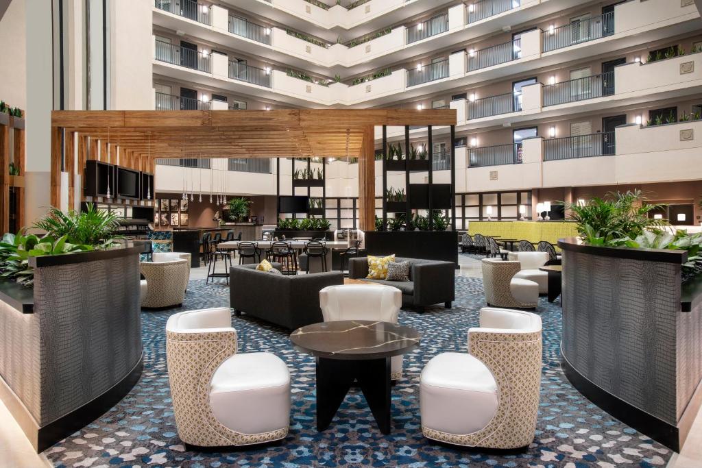 área comum do Embassy Suites Orlando - Airport com designs diferentes nas poltronas, estilo industrial com plantas e várias mesas com tampo de mármore e, atrás, há a fachada do hotel com janelinhas e varandas