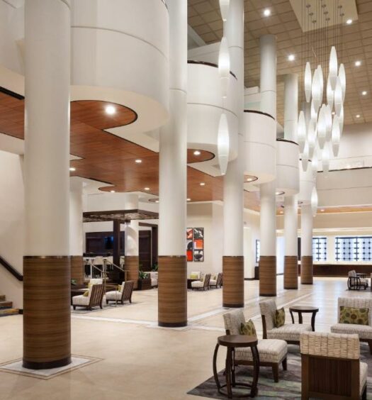 área comum do Marriott Orlando Airport Lakeside, um dos hotéis perto do aeroporto de Orlando, com colunas altas, escada, mesa com cadeiras de madeira com estofamento em tons de marrom e dourado, com lustres trabalhados acima ovalados e com a recepção com detalhes em vidro das janelas