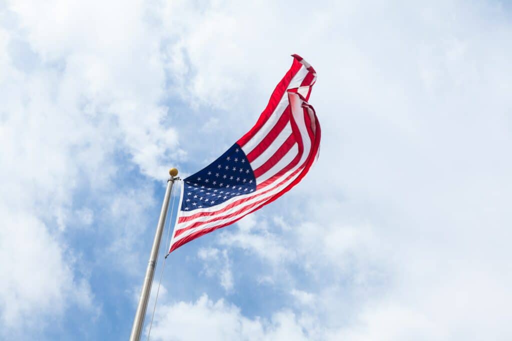 Bandeira dos EUA hasteada no topo de um mastro, com vento a deixando aberta, em um dia de céu repleto de nuvens, para ilustrar o post sobre como tirar visto americano
