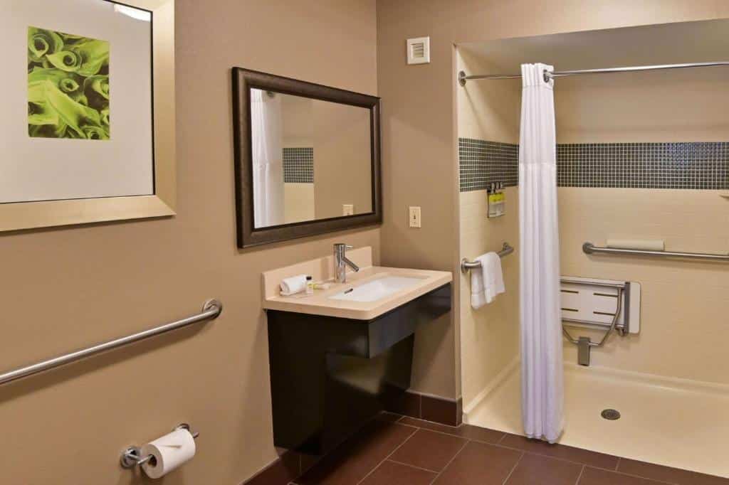 banheiro do Sonesta ES Suites Orlando - Lake Buena Vista com adaptações para PcDs, com pia aberta embaixo com espaço para cadeiras de rodas, há um box com cortina, chuveirinho, cadeira de banho e mais barras de apoio