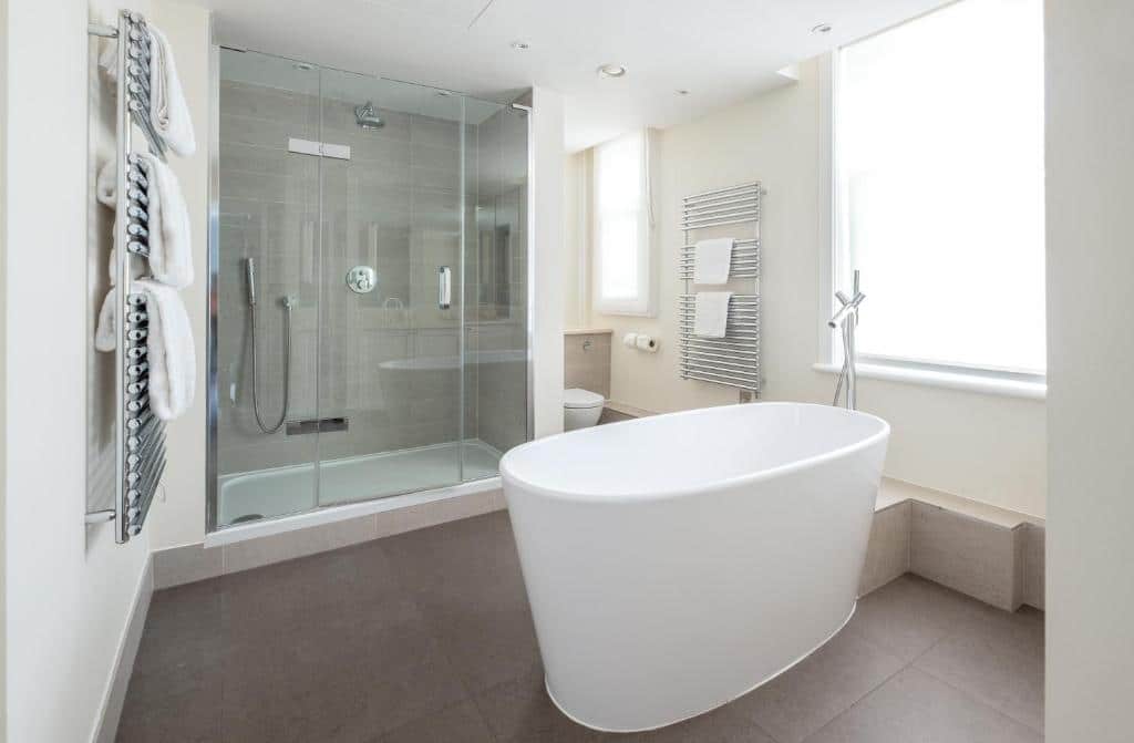 Banheiro do Native Mayfair com uma banheira oval, um box de vidro, prateleiras com toalhas brancas, para representar aluguel de temporada em Londres