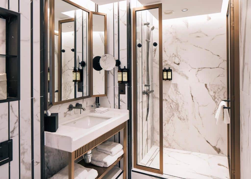 Banheiro do Page8, Page Hotels todo em mármore e com detalhes em cobre, box de vidro, pia quadrada e uma espelho amplo, há também prateleiras com toalhas brancas