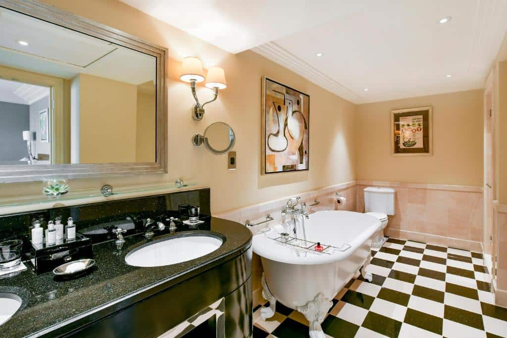 Banheiro amplo do The Savoy com uma banheira oval, um box, uma pia com duas cubas, uma espelho amplo e dois quadros nas paredes, para representar hotéis para família em Londres