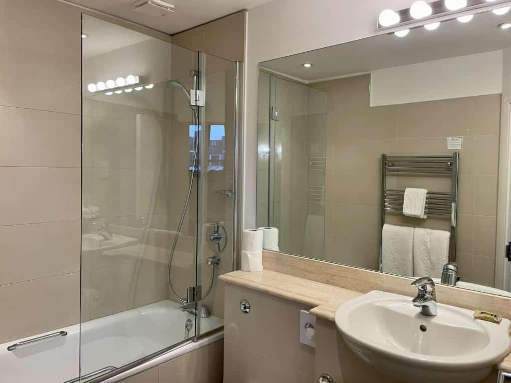 Banheiro do The Windermere Hotel, London com banheira de hidromassagem, uma pia e um espelho amplo, há também toalhas brancas penduradas pelo ambiente