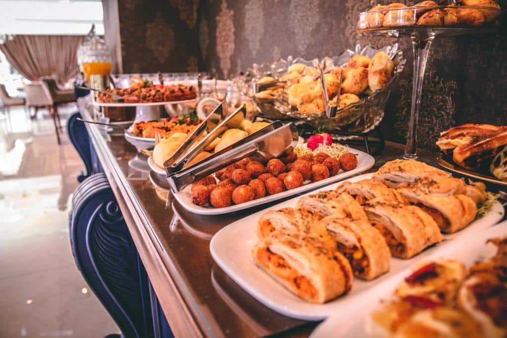 mesa de comidas do Hotel Daara com vários pratos estreitos e longos, repletos de alimentos, como folheados, salgados fritos, pães e bolos.