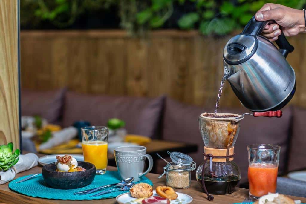 mesa de café da manhã do Wood Hotel com dois copos de vidro com sucos naturais, uma cumbuca de pedra com uma refeição elegante ovos e uma mão morena servindo um café recém coado.