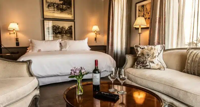 Quarto do The Singular Santiago com cama de casal ao fundo a frente uma mesa redonda com uma garrafa de vinho e duas taças e sofá ao lado da mesa.