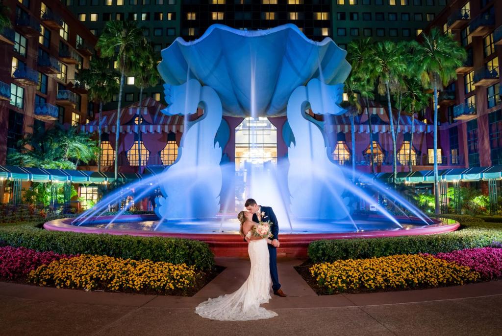 fonte estilizada com cisnes segurando uma concha, muitos jatos de água e tons de roxo e rosa em um jardim com um casal vestido de noivos se beijando no Walt Disney World Swan, um dos hotéis românticos em Orlando