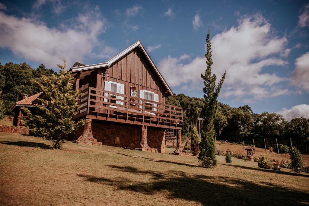Vist frontal do Chalés Pé da Serra em Gramado. Uma construção em madeira, com varanda aberta e duas janelas brancas. Atrás é possível ver diversas árvores e um céu azul com nuvens brancas