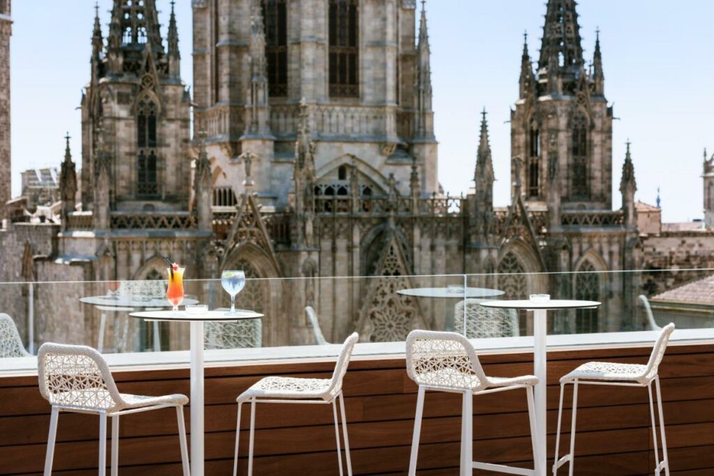 Terraço do Colón Hotel Barcelona, um dos melhores hotéis em Barcelona. Duas mesinhas brancas com cadeiras tem a Catedral de Barcelona atrás. Dois drinks estão em cima da mesa esquerda, e uma proteção de vidro está atrás das mesas.