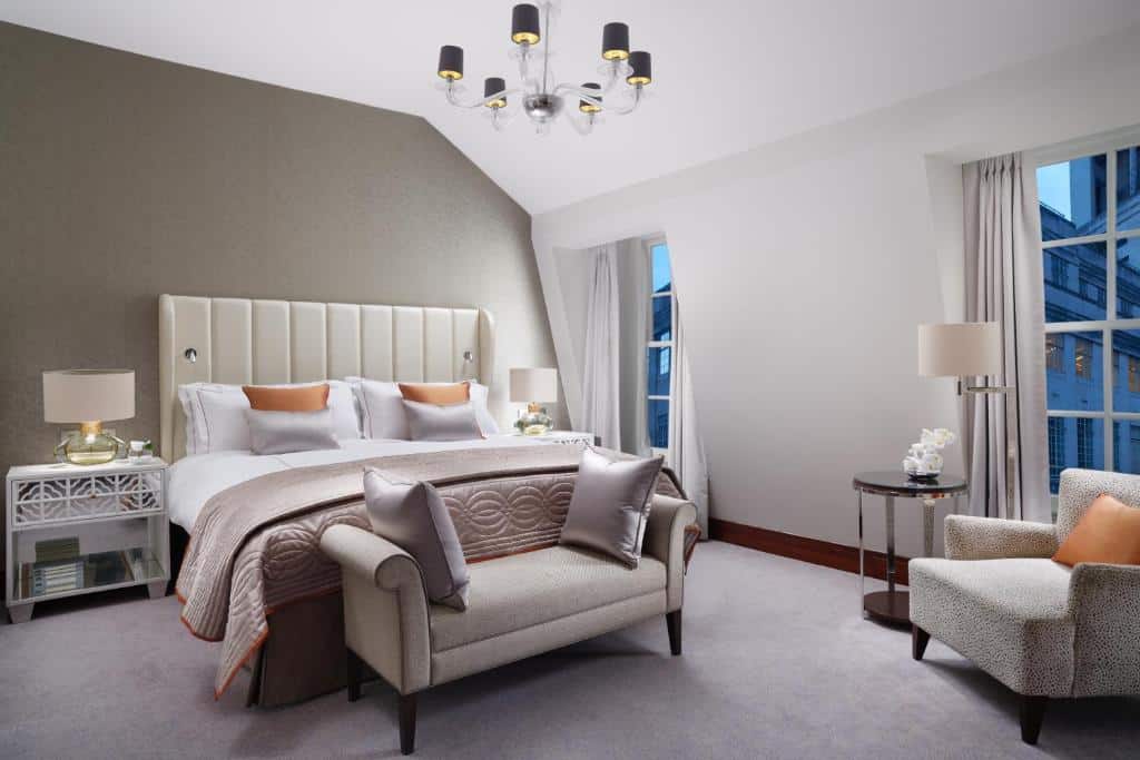 Quarto do Conrad London St James com duas janelas com cortinas, um carpete cinza, uma cama de casal, duas poltronas, uma mesinha de cabeceira com abajur e um lustre, para representar hotéis perto do Big Ben em Londres