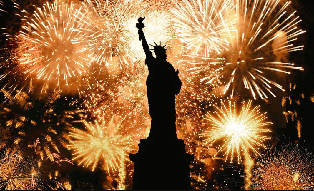 A Estátua da Liberdade, uma figura feminina romana segurando uma tocha em uma das mãos, com fogos de artíficio queimando ao fundo em tons de amarelo e dourado, para representar o réveillon em Nova York