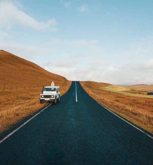 Estrada reta na Islândia, com vegetação cor de palha em ambos os lados da pista, e um carro branco parado um pouco atrás da metade visível da estrada, para ilustrar o guia definitivo sobre aluguel de carros para viagens