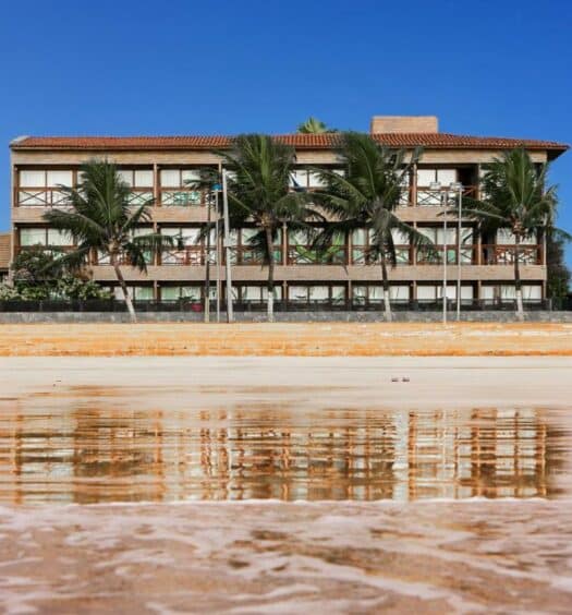 Vista da frente do Hotel Areias Belas, em Maragogi, sendo o local à beira-mar. Na imagem, é possível ver o mar, a areia e o hotel ao fundo com algumas árvores ao lado