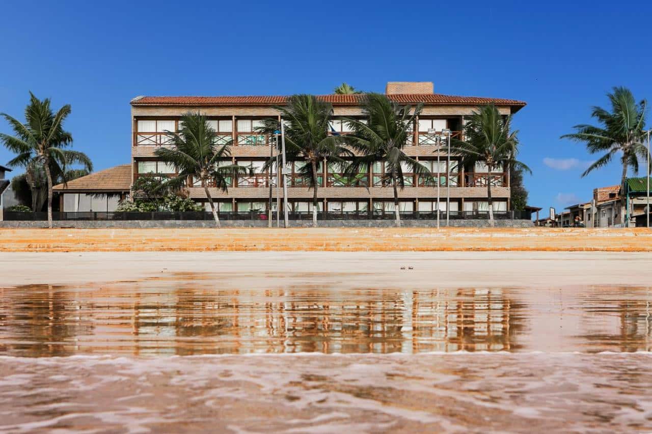 Vista da frente do Hotel Areias Belas, em Maragogi, sendo o local à beira-mar. Na imagem, é possível ver o mar, a areia e o hotel ao fundo com algumas árvores ao lado