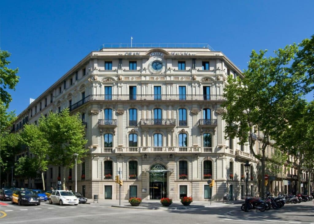 Fachada do Gran Hotel Havana, um dos melhores hotéis em Barcelona. O prédio é bege e tem cinco andares. Há motos e carros passando na rua ao redor, e algumas árvores e plantas na calçada.