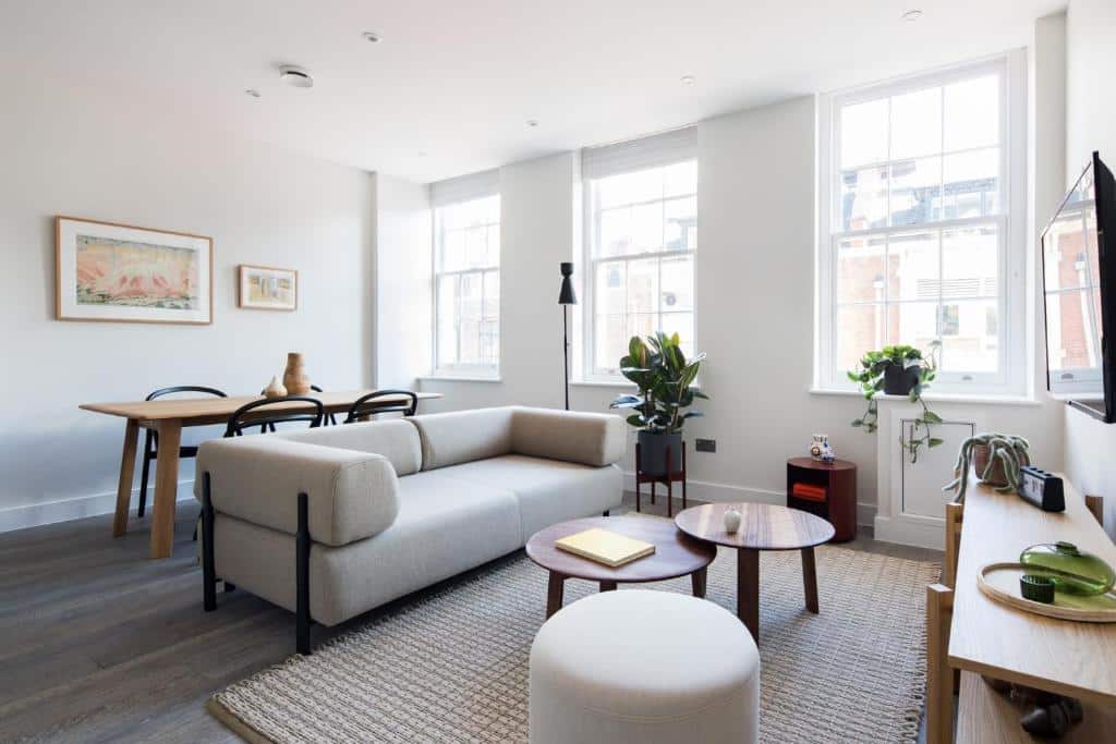 Sala de estar do apartamento Hausd – Leicester Square com uma sofá com dois lugares, três janelas, uma televisão, uma mesinha de centro redonda de madeira, um tapete bege, um bufê branco, uma mesa de quatro lugares e alguns vasos de plantas pelo ambiente