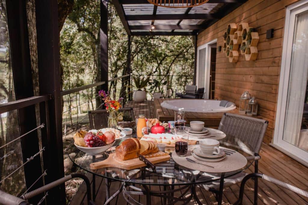varanda da Vila Nambu com uma mesa de vidro e ferro redonda repleta de alimentos para um café da manhã, três cadeira, uma banheira de hidromassagem redonda de dois lugares mais atrás e muitas árvores ao redor do chalé.